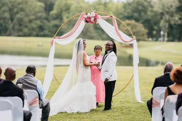 A couple exchanging vows under a floral arch on a grassy lawn near a lake, with guests seated in white chairs.