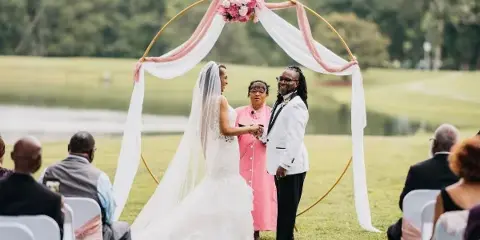 A couple exchanging vows under a floral arch on a grassy lawn near a lake, with guests seated in white chairs.