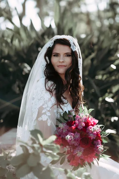 A bride with styled wavy hair and a lace veil holding a bouquet of pink and red flowers.
