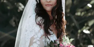 A bride with styled wavy hair and a lace veil holding a bouquet of pink and red flowers.