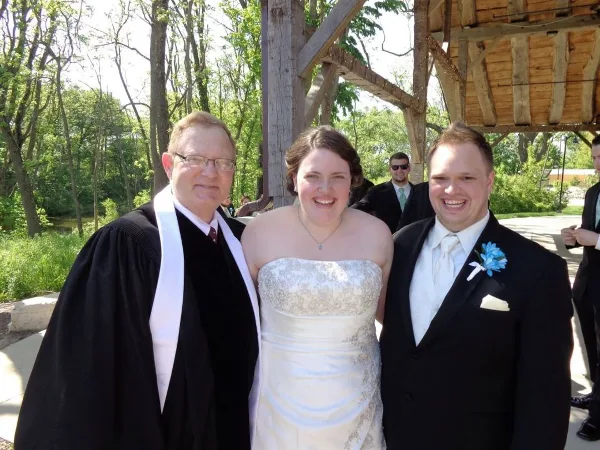A wedding officiant in black robes stands smiling with a bride and groom at an outdoor ceremony.