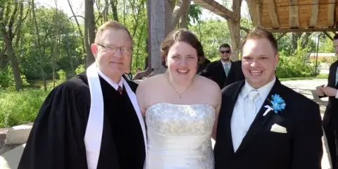 A wedding officiant in black robes stands smiling with a bride and groom at an outdoor ceremony.