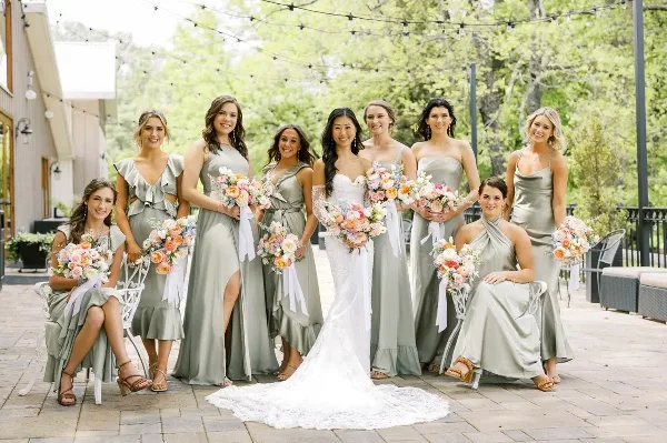 A bride and bridesmaids in sage green dresses holding floral bouquets at an outdoor wedding venue.