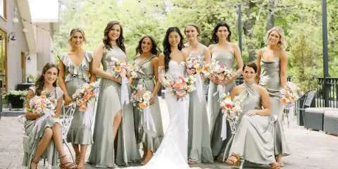 A bride and bridesmaids in sage green dresses holding floral bouquets at an outdoor wedding venue.