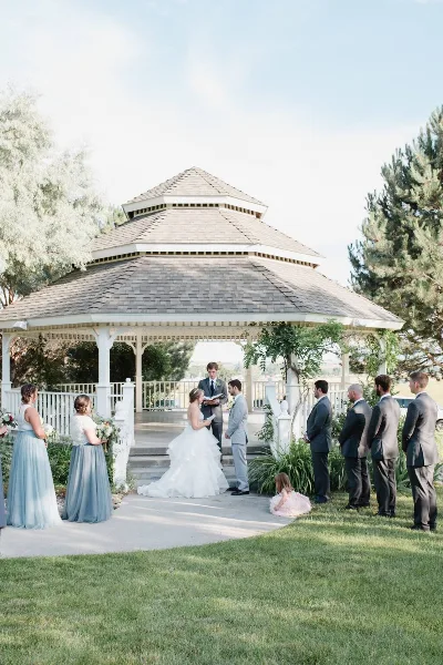 A wedding ceremony in progress with a bride and groom standing before an officiant inside a white, multi-tiered gazebo surrounded by a wedding party.