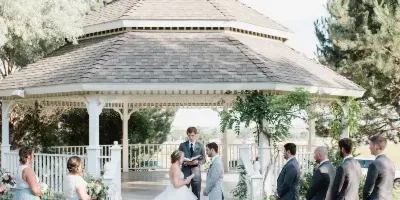A wedding ceremony in progress with a bride and groom standing before an officiant inside a white, multi-tiered gazebo surrounded by a wedding party.
