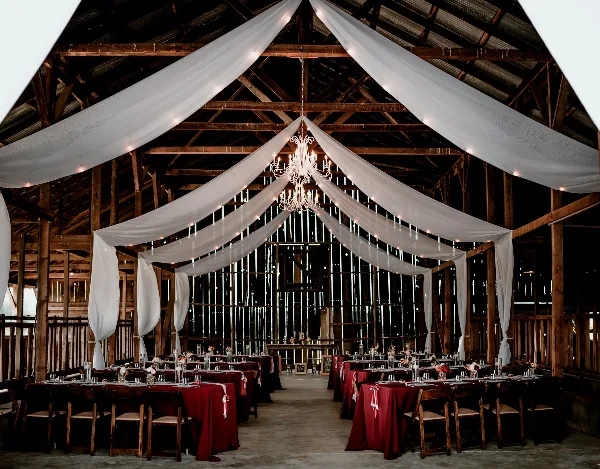 Interior of a rustic wooden barn venue decorated with white drapery, a chandelier, and tables set for a wedding reception.