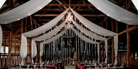 Interior of a rustic wooden barn venue decorated with white drapery, a chandelier, and tables set for a wedding reception.