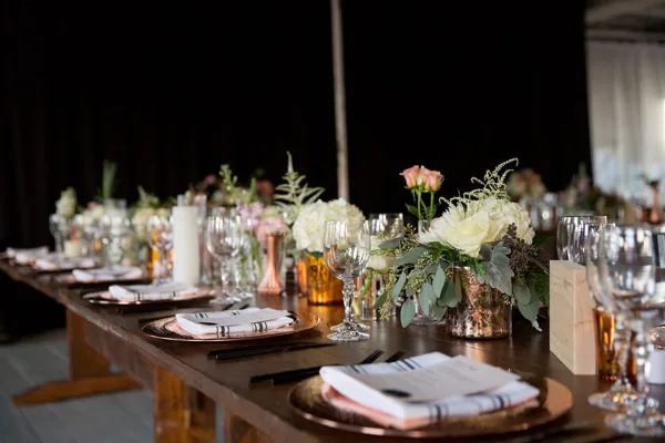 A long wooden table set for a wedding reception featuring white floral arrangements, candles, and copper accents.