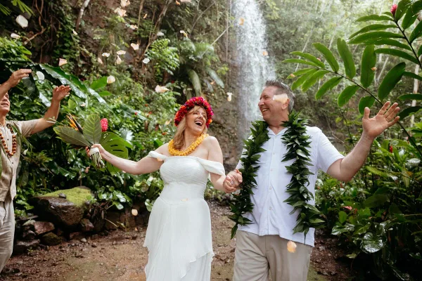 A bride and groom wearing Hawaiian leis and flower crowns celebrate with falling petals in front of a tropical waterfall.