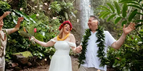 A bride and groom wearing Hawaiian leis and flower crowns celebrate with falling petals in front of a tropical waterfall.