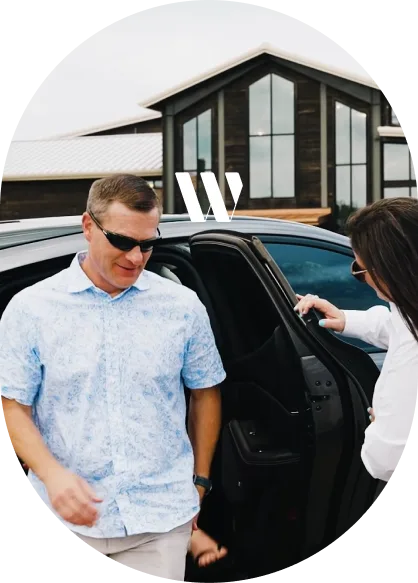 A man in a light blue shirt exiting a black luxury sedan while a woman holds the door open.