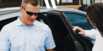 A man in a light blue shirt exiting a black luxury sedan while a woman holds the door open.