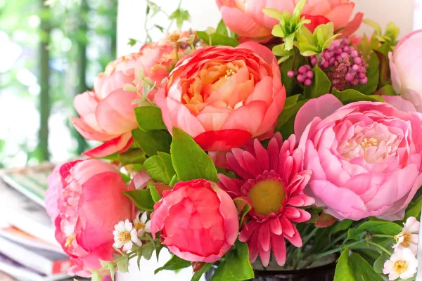 A vibrant floral arrangement featuring pink and coral peonies, a pink daisy, and green foliage.