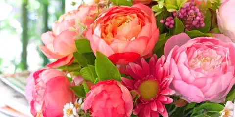 A vibrant floral arrangement featuring pink and coral peonies, a pink daisy, and green foliage.