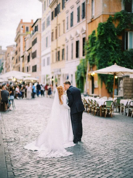 A bride and groom kissing on a cobblestone street with outdoor cafe seating in the background.
