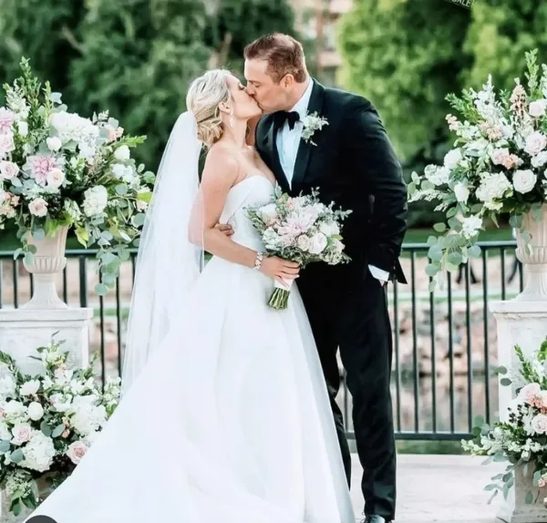 A bride and groom kissing, with the bride holding a large bouquet of white and pink flowers, flanked by tall floral arrangements in urns.