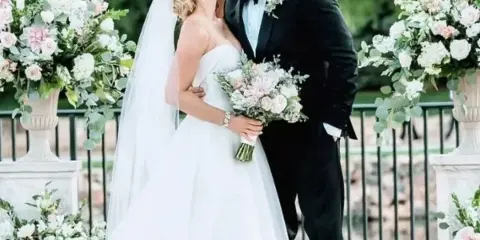 A bride and groom kissing, with the bride holding a large bouquet of white and pink flowers, flanked by tall floral arrangements in urns.