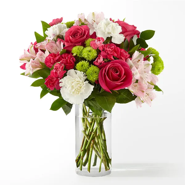 A floral arrangement featuring pink roses, white and pink carnations, and green button chrysanthemums in a clear cylindrical glass vase.