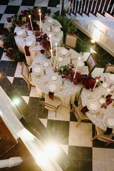 Overhead view of an elegant wedding reception table setting featuring white linens, red floral arrangements, and tall candles on a checkered floor.