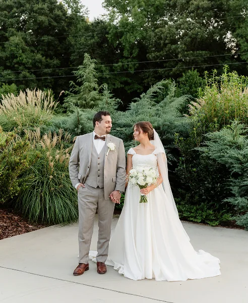 A bride and groom standing together outdoors in a garden setting, holding hands and looking at each other.