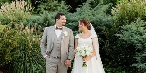 A bride and groom standing together outdoors in a garden setting, holding hands and looking at each other.