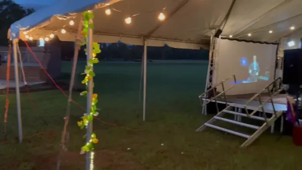 An outdoor event setup featuring a large white tent with string lights, a stage with stairs, and a projection screen.