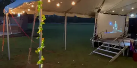 An outdoor event setup featuring a large white tent with string lights, a stage with stairs, and a projection screen.