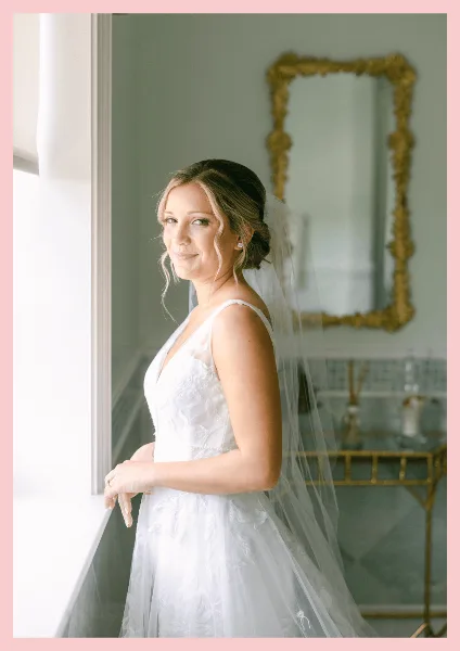 A bride in a white wedding dress and veil standing by a window with styled hair and makeup.