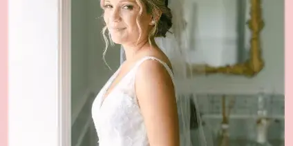 A bride in a white wedding dress and veil standing by a window with styled hair and makeup.