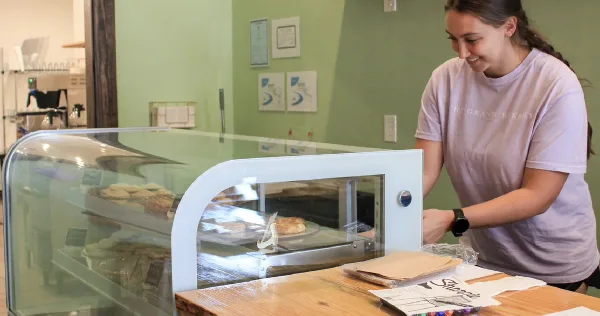 Bakery staff member in a branded shirt serving customers at a glass display case filled with baked goods.