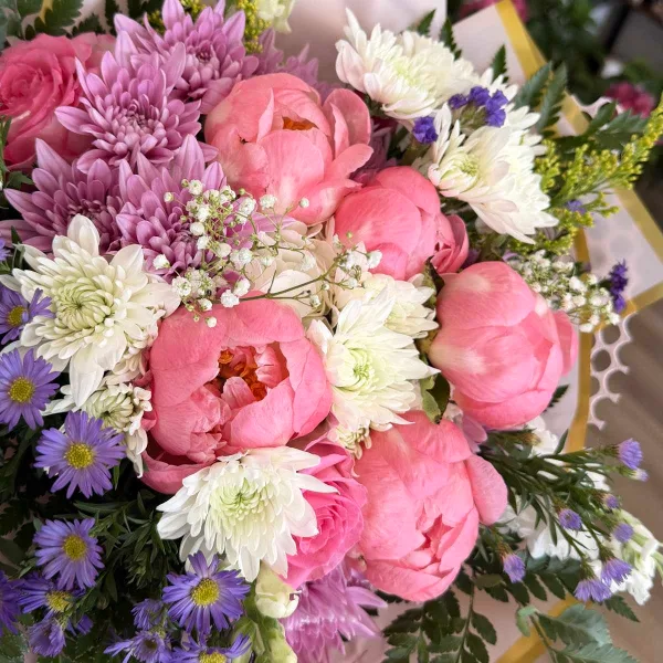 A close-up of a floral arrangement featuring pink peonies, white and purple chrysanthemums, and purple asters.