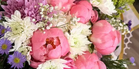 A close-up of a floral arrangement featuring pink peonies, white and purple chrysanthemums, and purple asters.