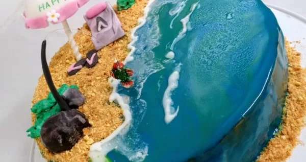 A custom themed cake featuring a beach scene with a blue fondant ocean, sandy beach, and decorative elements.