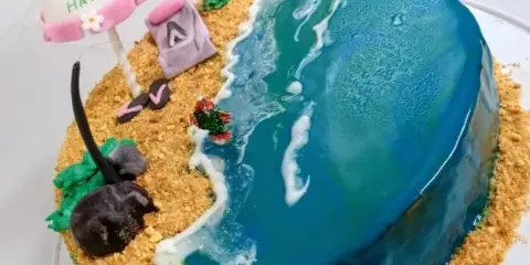 A custom themed cake featuring a beach scene with a blue fondant ocean, sandy beach, and decorative elements.