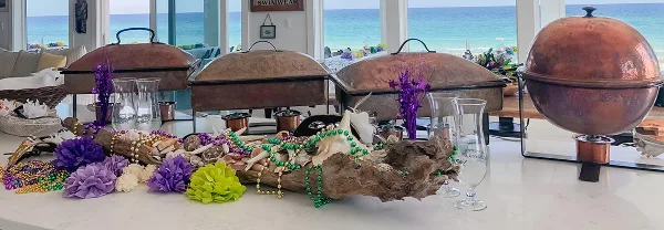 A catering buffet setup featuring copper chafing dishes, purple and green floral decorations, and a view of the ocean.