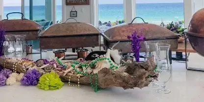 A catering buffet setup featuring copper chafing dishes, purple and green floral decorations, and a view of the ocean.