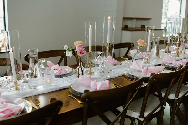 A professionally arranged wedding reception table featuring white plates, pink napkins, tall glass candle holders, and floral centerpieces.