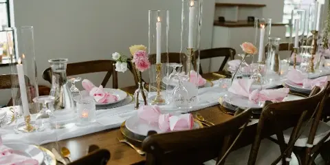 A professionally arranged wedding reception table featuring white plates, pink napkins, tall glass candle holders, and floral centerpieces.