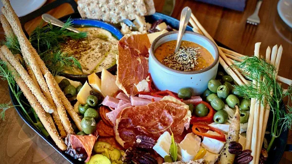 A large, artfully arranged charcuterie board featuring cured meats, assorted cheeses, green olives, breadsticks, and a dipping sauce, garnished with fresh dill.