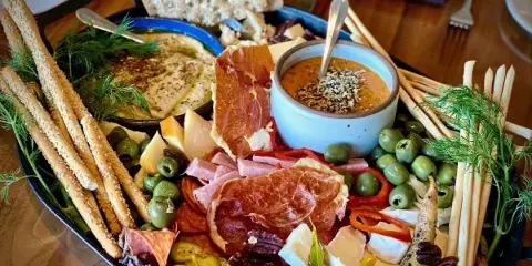 A large, artfully arranged charcuterie board featuring cured meats, assorted cheeses, green olives, breadsticks, and a dipping sauce, garnished with fresh dill.