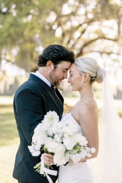 A professional wedding photograph of a bride and groom standing close together outdoors, with the groom in a dark suit and the bride in a white dress holding a bouquet of white flowers.