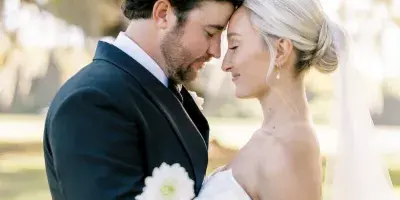 A professional wedding photograph of a bride and groom standing close together outdoors, with the groom in a dark suit and the bride in a white dress holding a bouquet of white flowers.