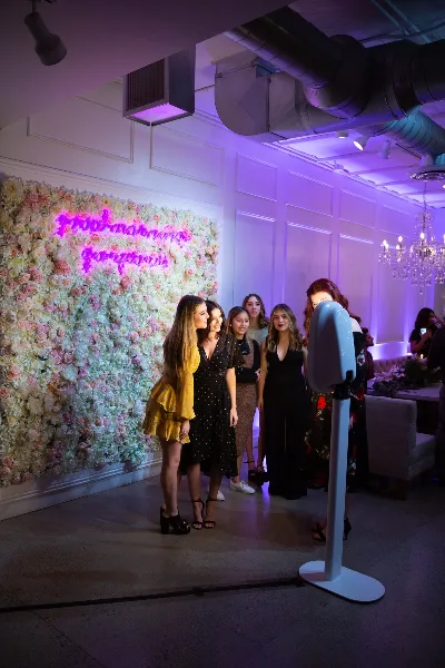 A group of women posing in front of a floral wall and a standing photo booth at a wedding reception.