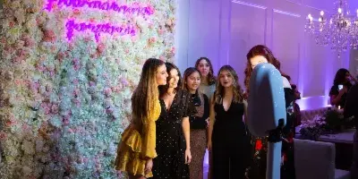 A group of women posing in front of a floral wall and a standing photo booth at a wedding reception.