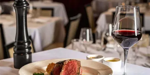 A plated steak dish with a glass of red wine on a restaurant table setting.