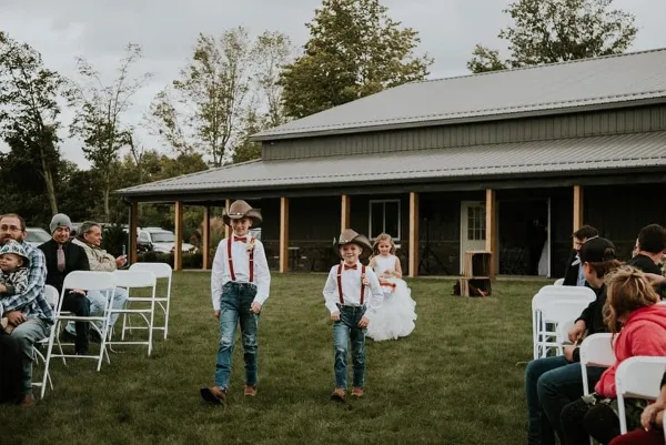 Outdoor wedding ceremony at a barn venue with flower girls walking down the aisle.
