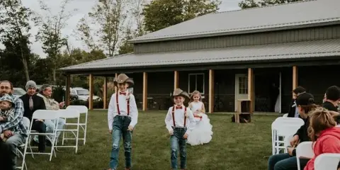 Outdoor wedding ceremony at a barn venue with flower girls walking down the aisle.