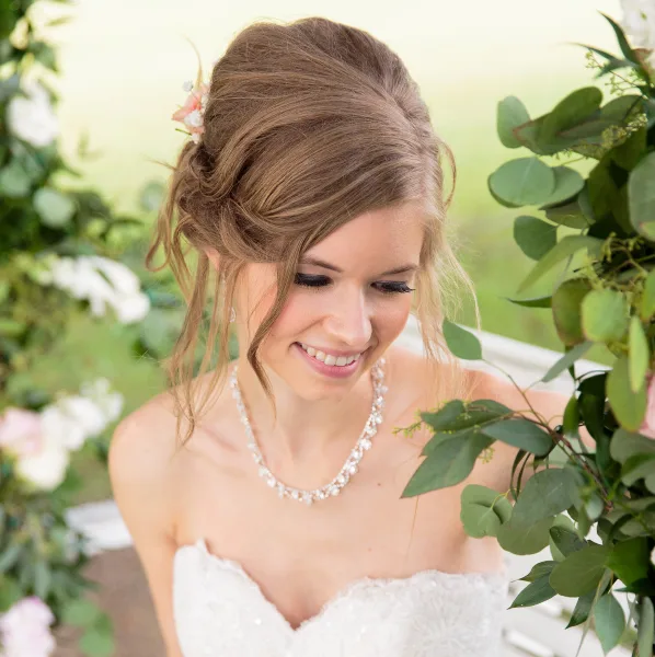 A smiling bride with an elegant updo hairstyle and professional makeup, wearing a white wedding dress and pearl necklace, standing near a floral arch.