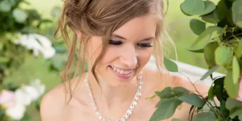 A smiling bride with an elegant updo hairstyle and professional makeup, wearing a white wedding dress and pearl necklace, standing near a floral arch.
